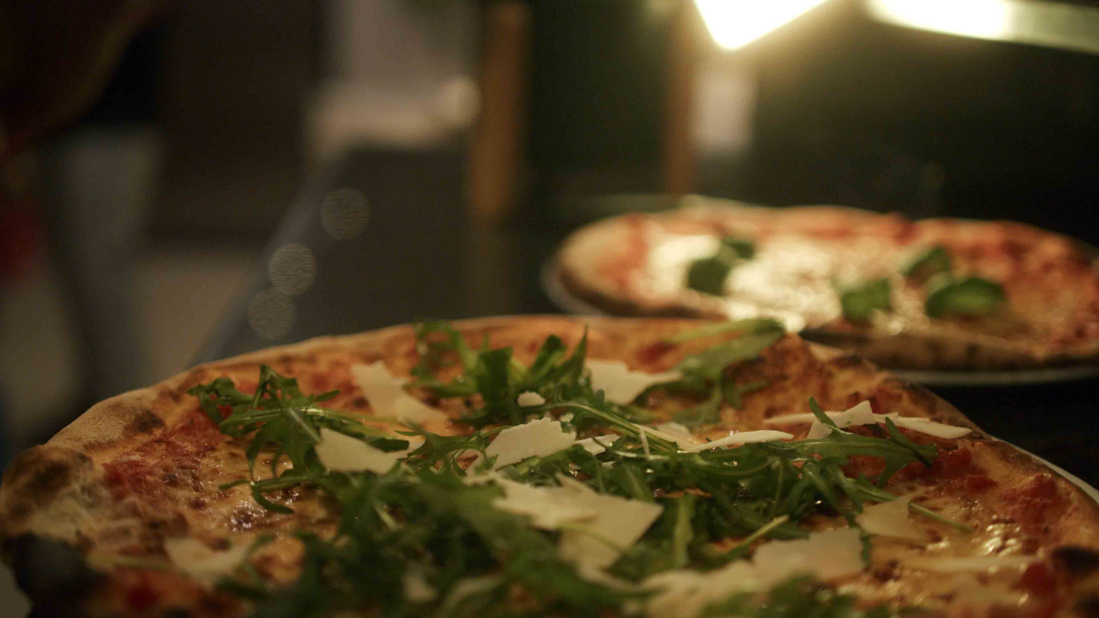 Zwei Pizzen auf Tellern, eine davon mit Rucola und gehobeltem Käse, in weichem, warmem Licht.